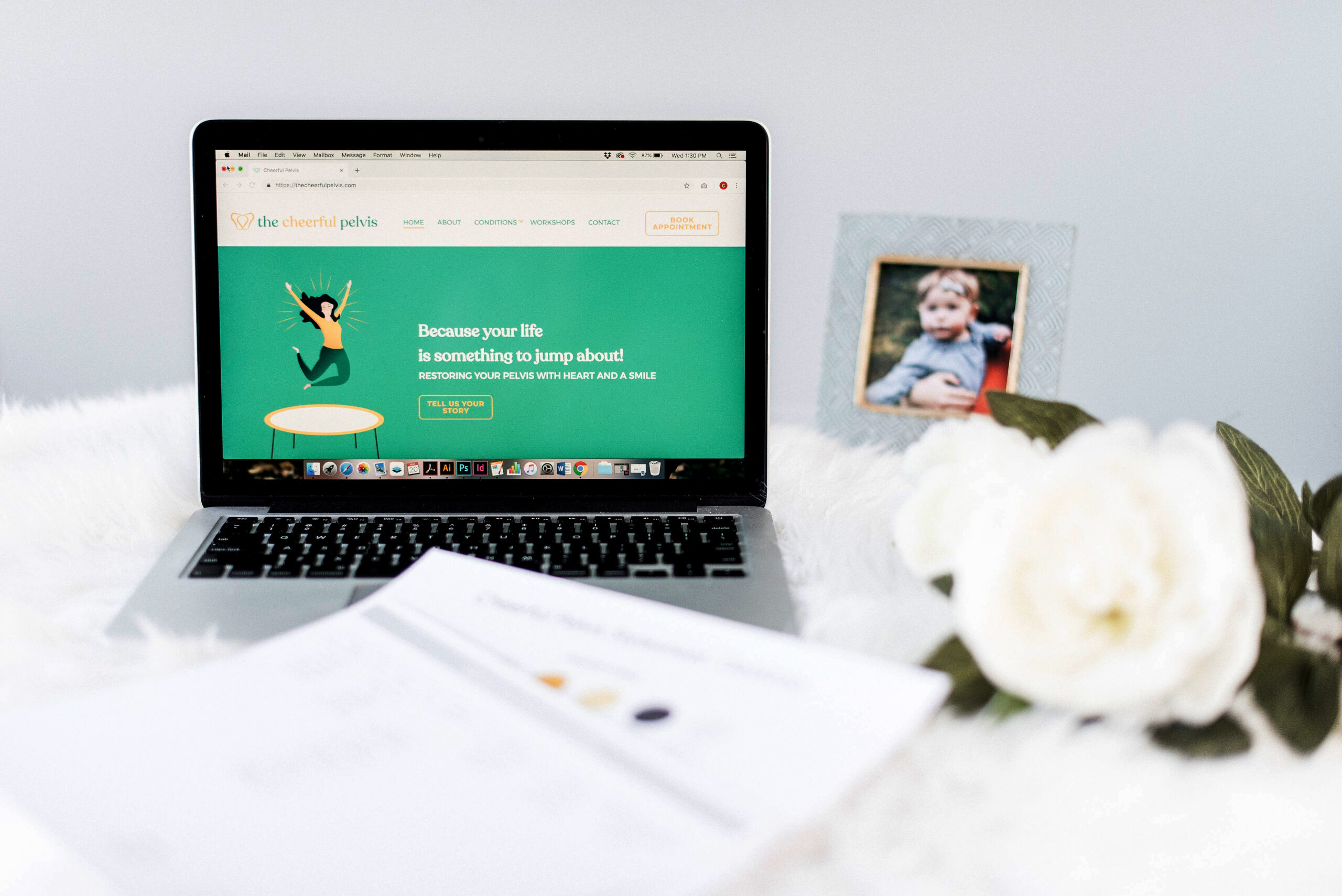 Website for the Cheerful Pelvis on a laptop sitting next to flowers and a photograph of a child